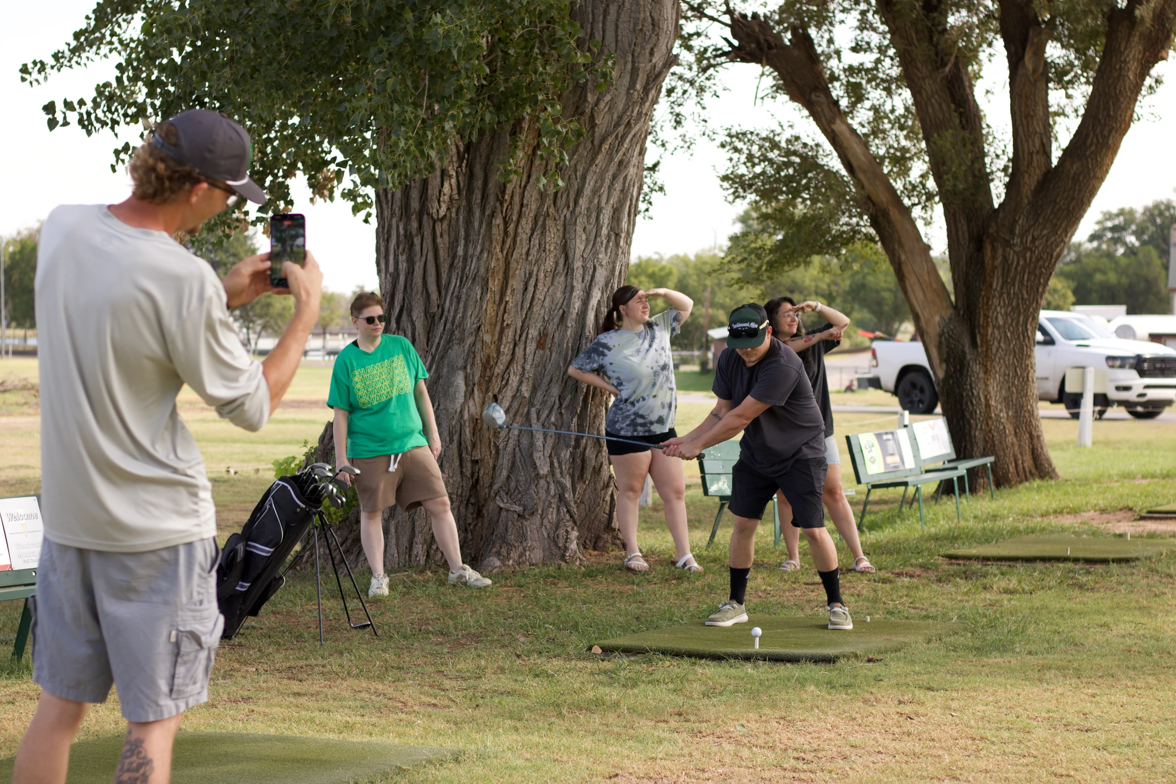 A tech captain films a teammate’s golf swing beneath a large tree.